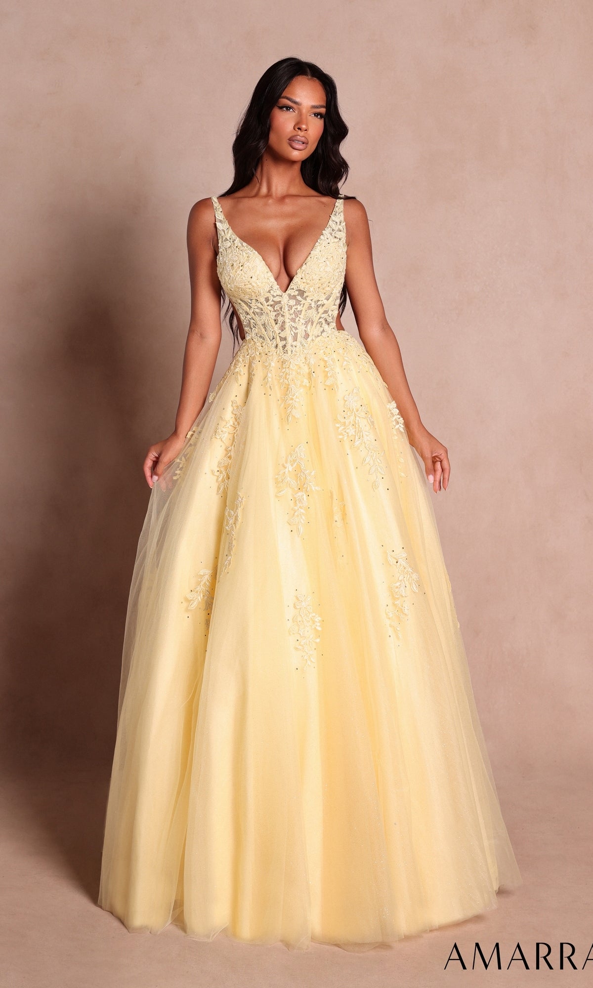 A woman wearing the Amarra 89786 long prom dress in light yellow, featuring a fitted beaded bodice with delicate floral lace appliqués and a flowing tulle skirt, stands against a plain beige backdrop.